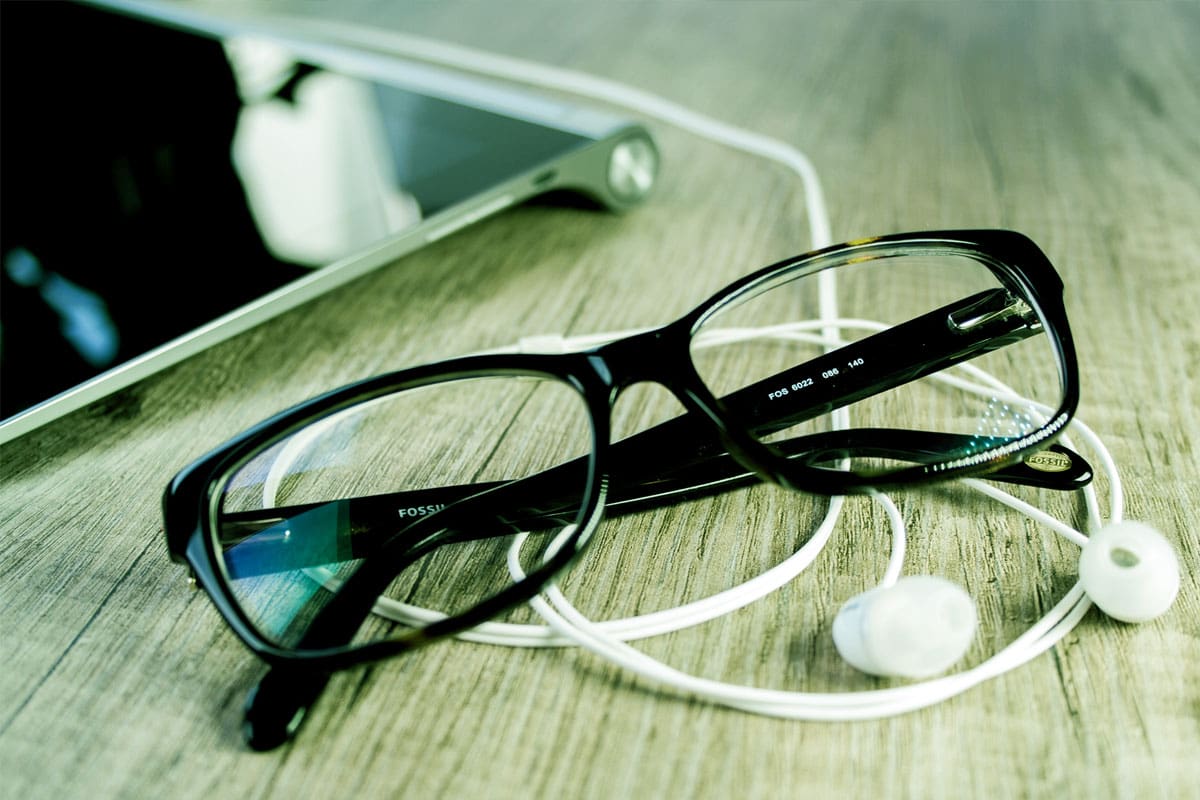 Eyeglasses, wired earbuds, and a tablet on a wooden surface.