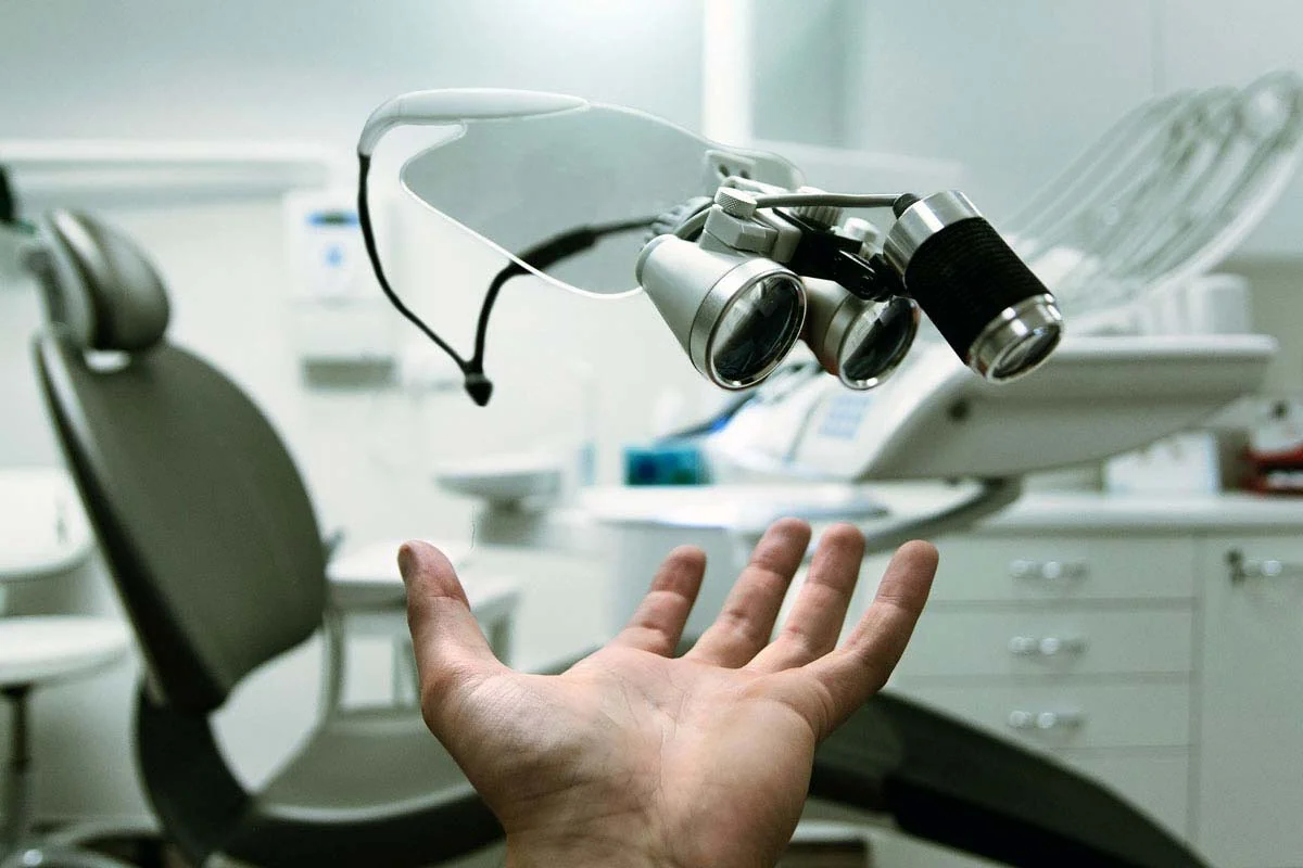 A hand reaching up towards a pair of dental loupes in a dental office.